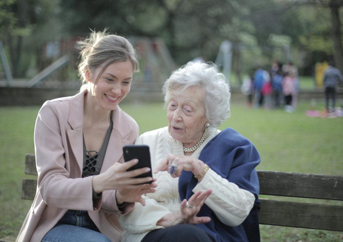 Delighted female relatives sitting together on wooden bench in park and browsing mobile phone while learning using